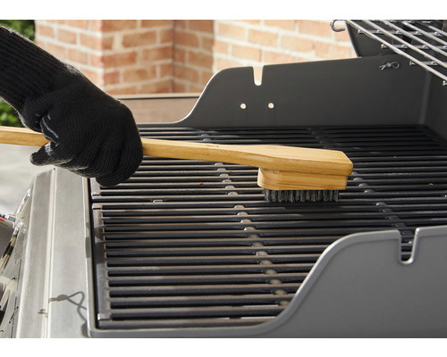 Een hand met een barbecuehandschoen reinigt een grillrooster met een barbecueborstel met houten handvat.