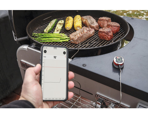 Foto van een barbecue met steaks, maïskolven, courgette en asperges, waarbij een smartphone de temperatuur weergeeft.