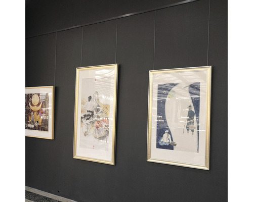 Drie ingelijste kunstwerken hangen aan een grijze muur, bevestigd met touwen