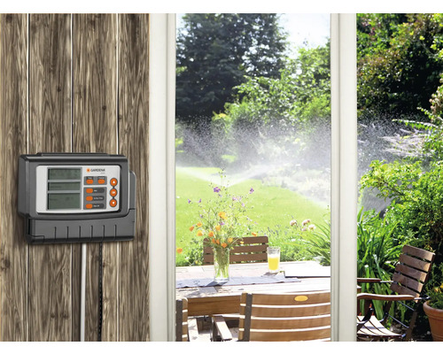 Gardena irrigatiecomputer voor tuinirrigatie gemonteerd op een houten muur. Op de achtergrond een tuin met gazonsproeier en een tuintafel.