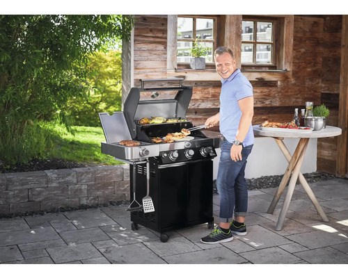 Scene met een man die vlees en groenten grilt op een zwarte gasbarbecue, buiten op een terras.