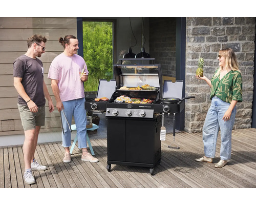 Drie mensen rond een gasbarbecue op een terras met gegrilde ingrediënten, koekenpan en ananas