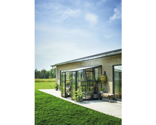 Een glazen serre met planten staat tegen een muur van een huis op een terras.