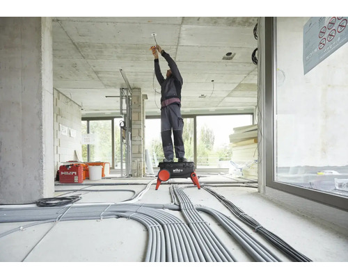 Een elektricien staat op een opstapje en werkt in een ruwbouw aan het plafond. Er liggen veel kabels op de grond.