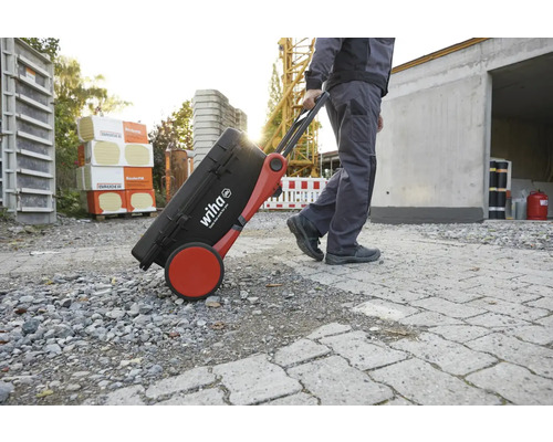 Een persoon trekt een zwarte Wiha gereedschapskoffer met rode wielen over een oneffen oppervlak.